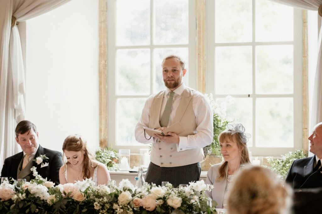 A groom giving a speech at a formal indoor wedding celebration with family and floral decorations.