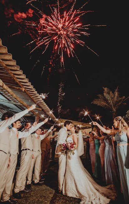 Bride and groom kiss under fireworks surrounded by wedding guests holding sparklers.